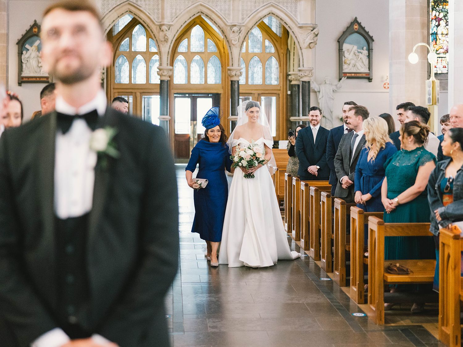 A wedding ceremony takes place inside a beautiful gothic church with stained glass windows and wooden pews lined with guests.