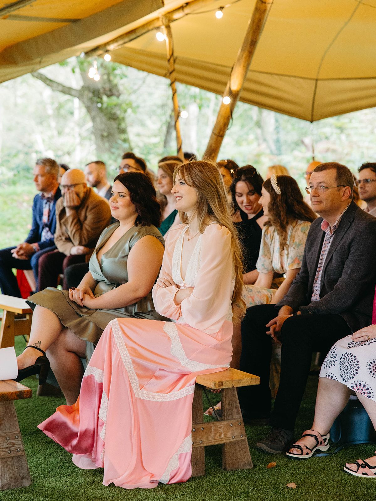 Guests seated on wooden benches under a tan canopy tent watching an outdoor event.