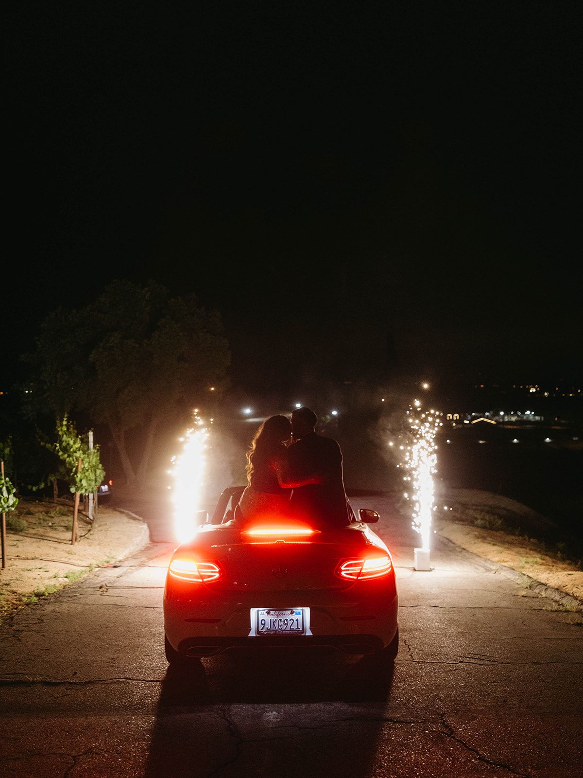 Red car parked at night with sparklers creating light trails on both sides, taillights glowing against dark background.