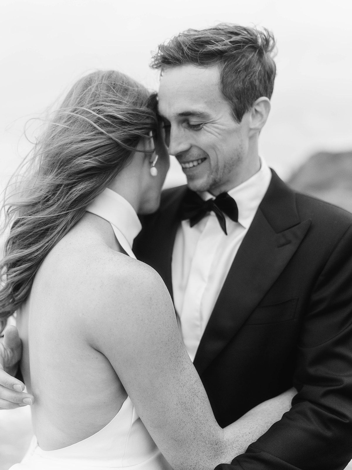 A romantic black and white portrait of a couple in wedding attire embracing by the sea.