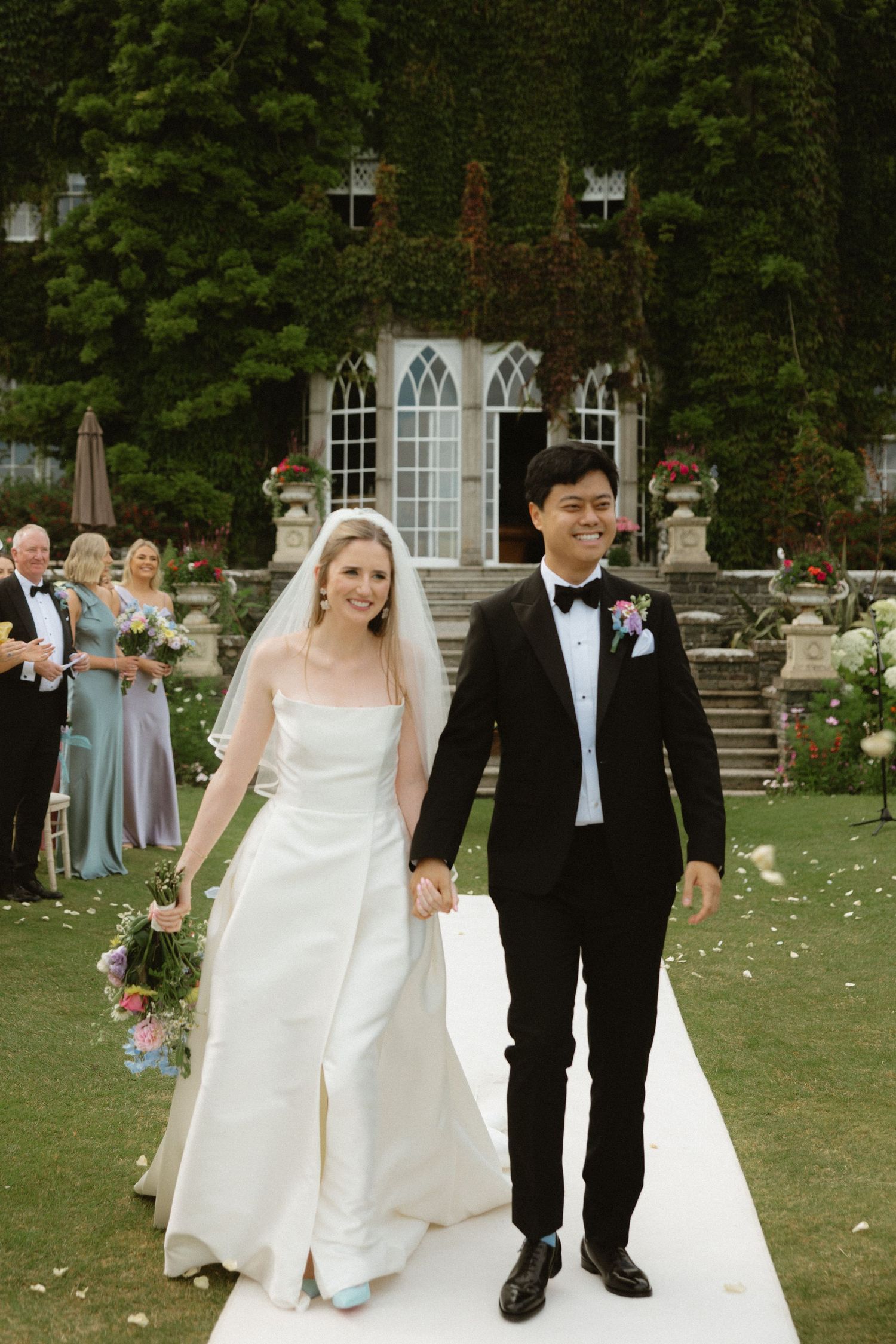 Newlyweds walk down white aisle runner after outdoor ceremony in front of ivy-covered building.