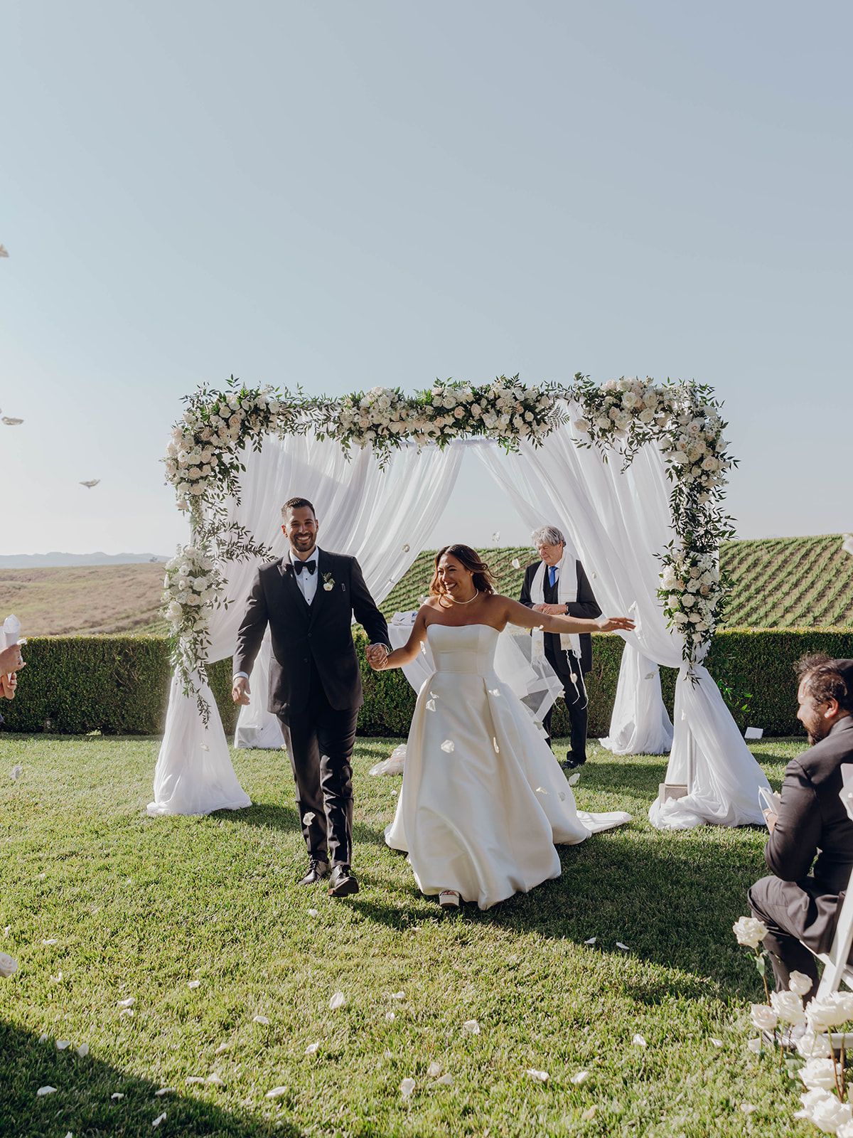A joyful wedding celebration set outdoors with a floral arch canopy, white drapes, and vineyard views in the background.