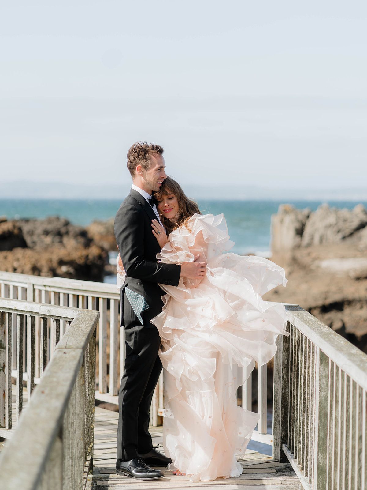 Romantic moment between newlyweds on oceanfront boardwalk with dress flowing in wind.