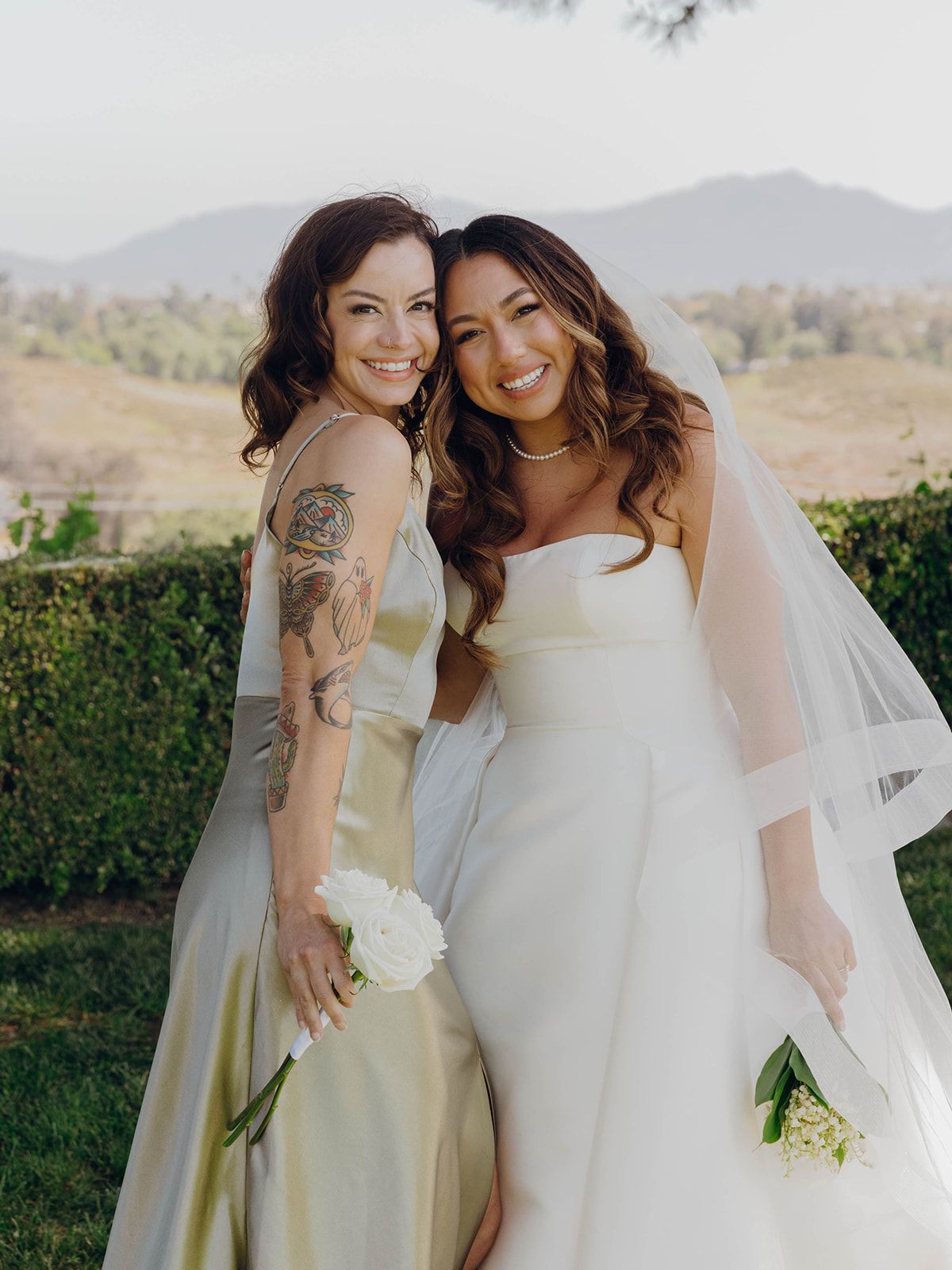 Two people in formal attire embrace and smile outdoors at a desert wedding with mountains in the background.