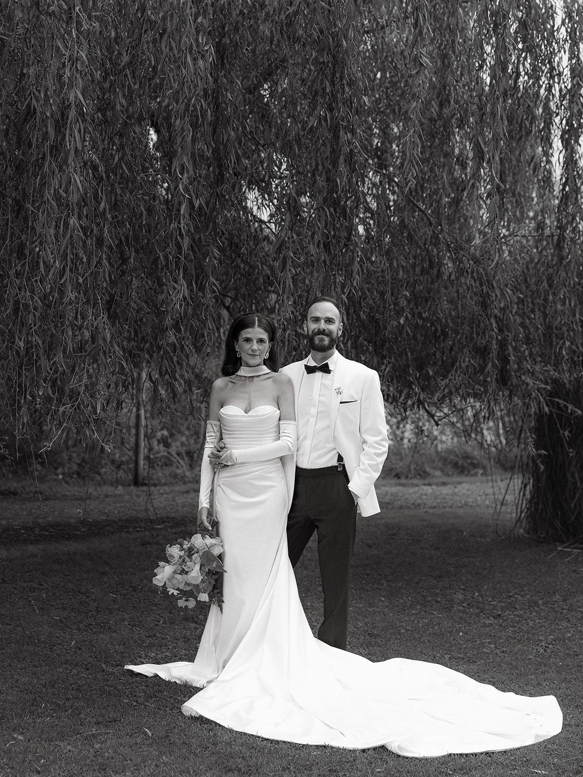 Black and white wedding portrait showing a couple posing beneath drooping willow trees.