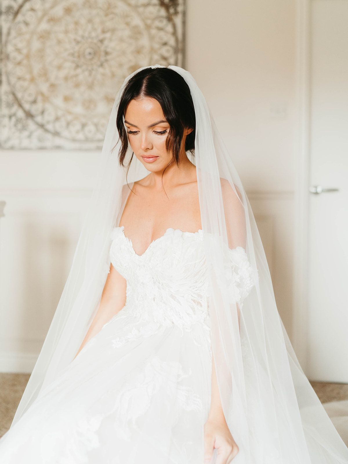 A bride in a flowing white wedding dress and veil stands gracefully in a bright, elegant room with patterned wallpaper.