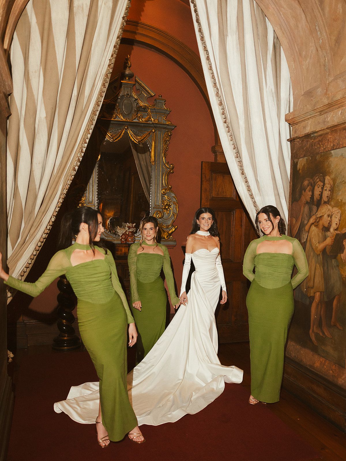 A wedding party in green bridesmaid dresses and white bridal gown pose together in an elegant indoor venue.