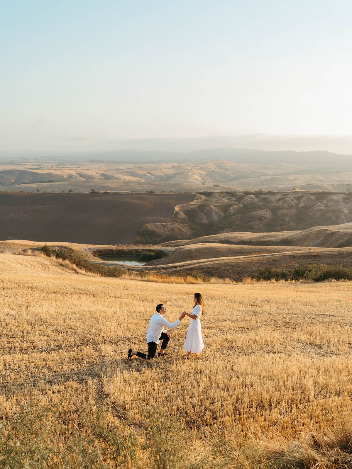 Two figures sit together in a golden field overlooking rolling hills at sunset.