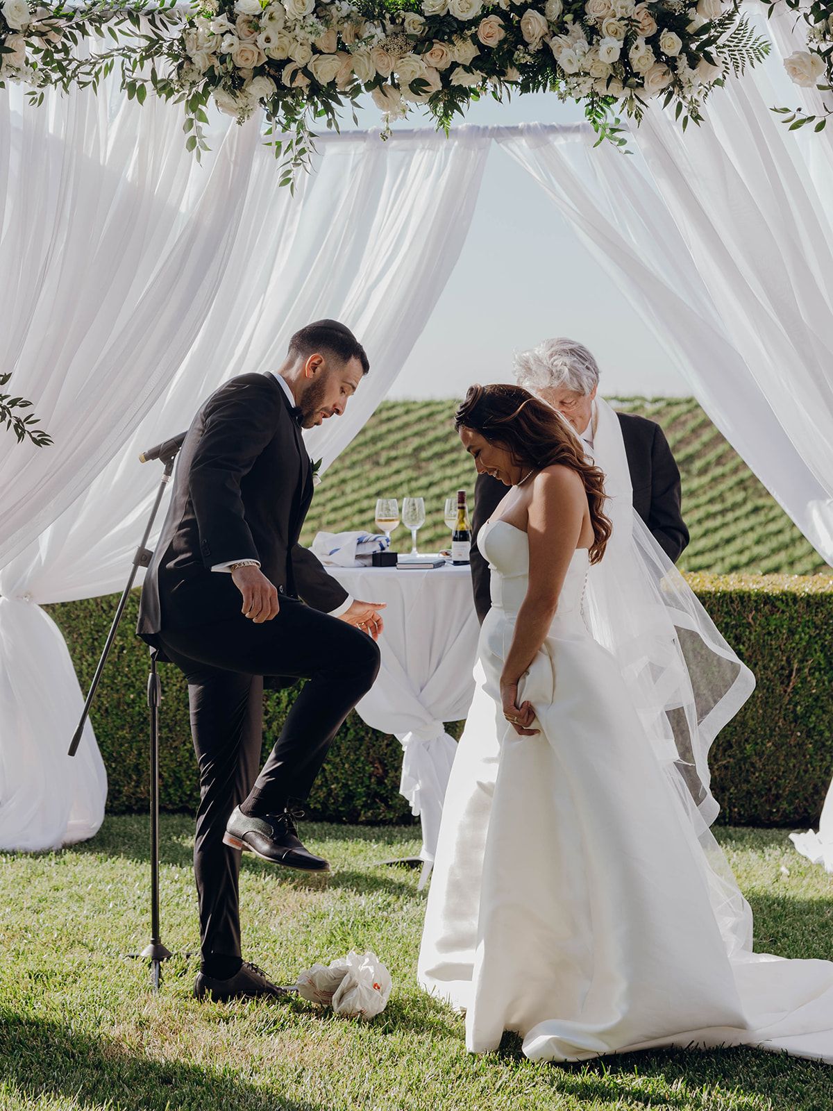A couple shares a playful moment with a soccer ball under an elegant wedding arch with white drapes and greenery outdoors.