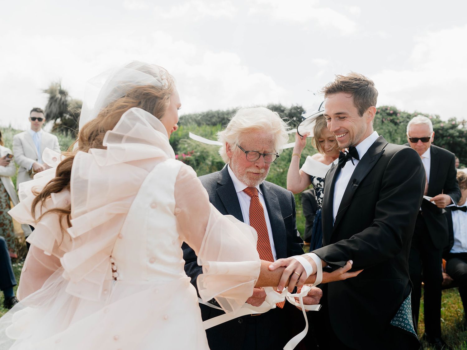 Couple exchanges rings during outdoor wedding ceremony with officiant in formal attire.