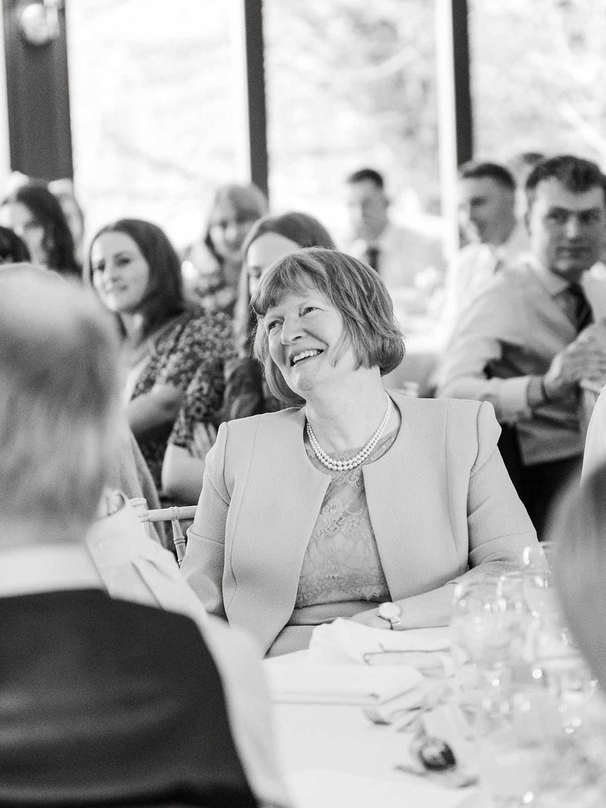 A joyous guest in a light-colored jacket and pearl necklace laughs heartily during a formal dining event.
