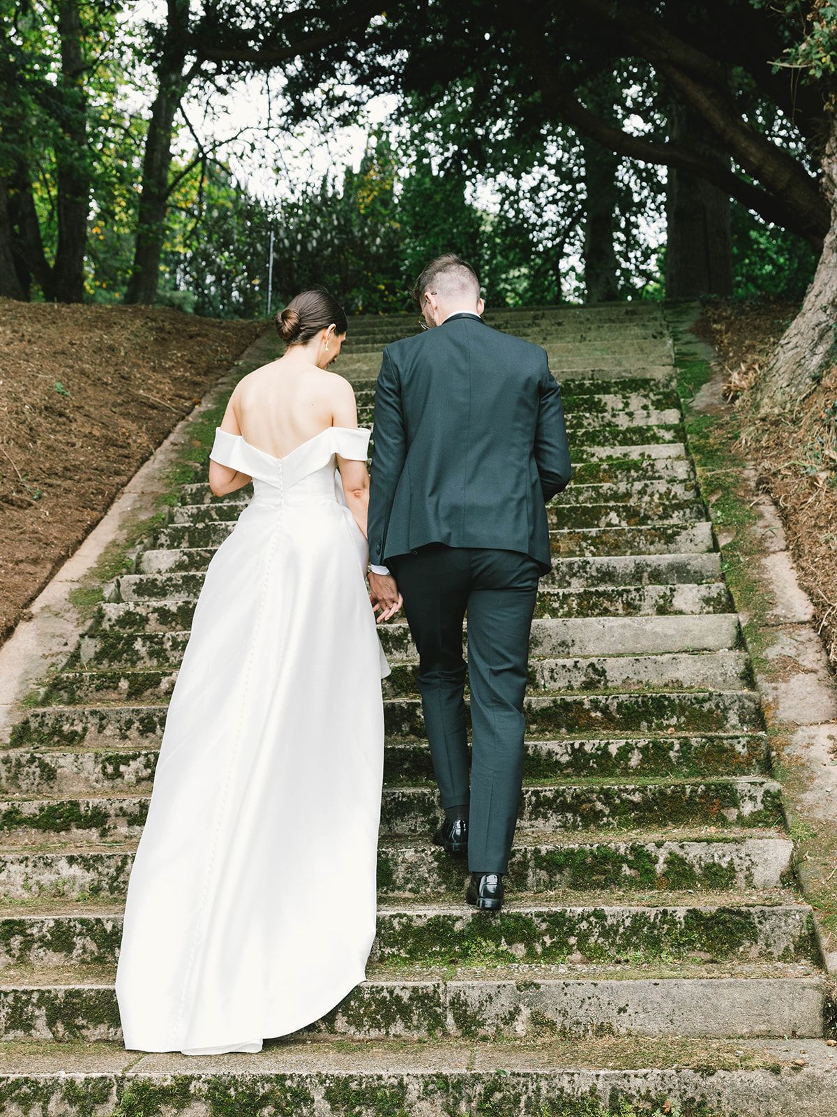 A couple in wedding attire walking up rustic stone steps surrounded by lush green trees and foliage.