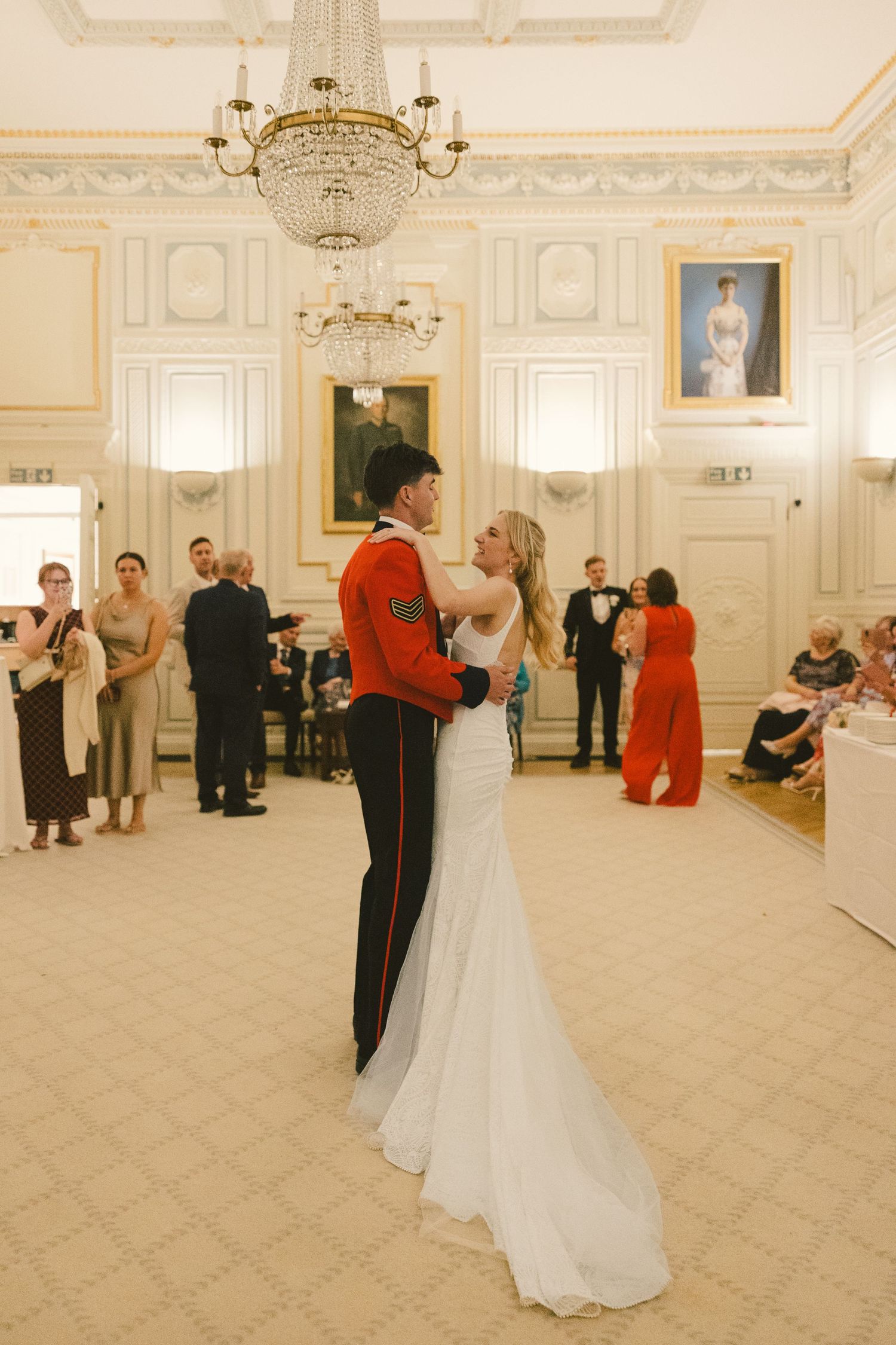 A couple shares their first dance in an elegant ballroom with chandeliers and white walls as guests watch.