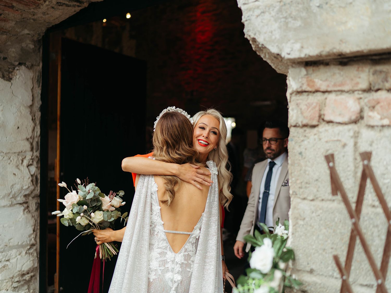 Two people share an emotional embrace in formal white attire outside a rustic stone building entrance.