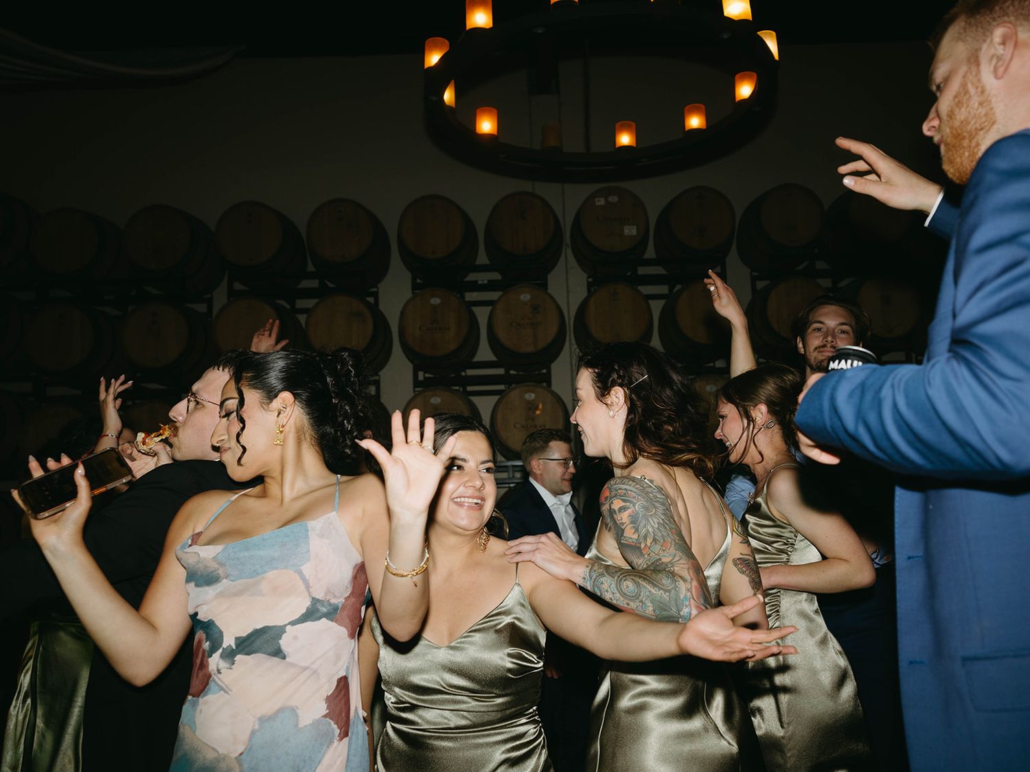 Party guests dance and celebrate in a dimly lit wine cellar with wooden barrels lining the walls.