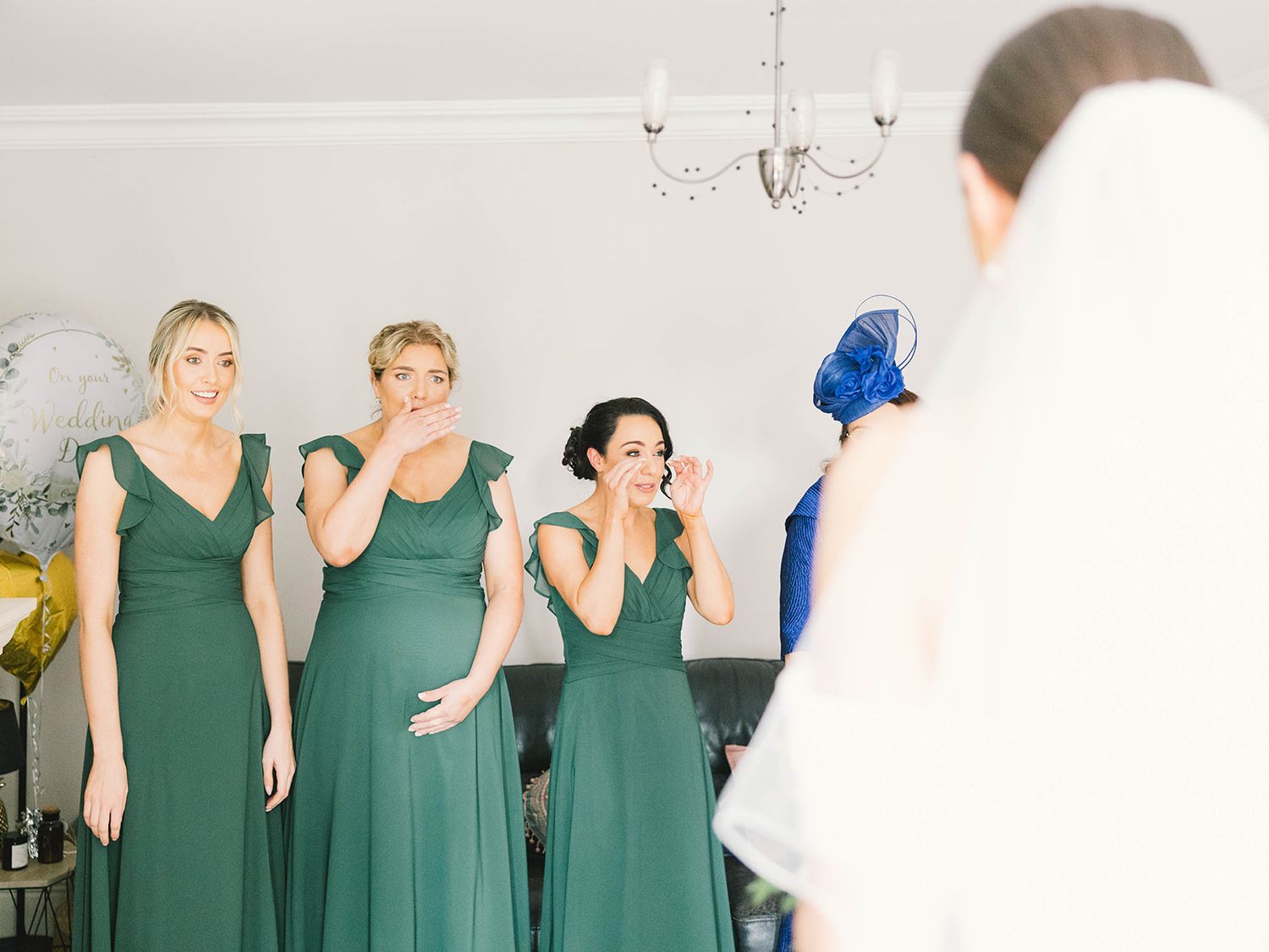 Three women in sage green bridesmaid dresses react emotionally to seeing someone in a white wedding dress.