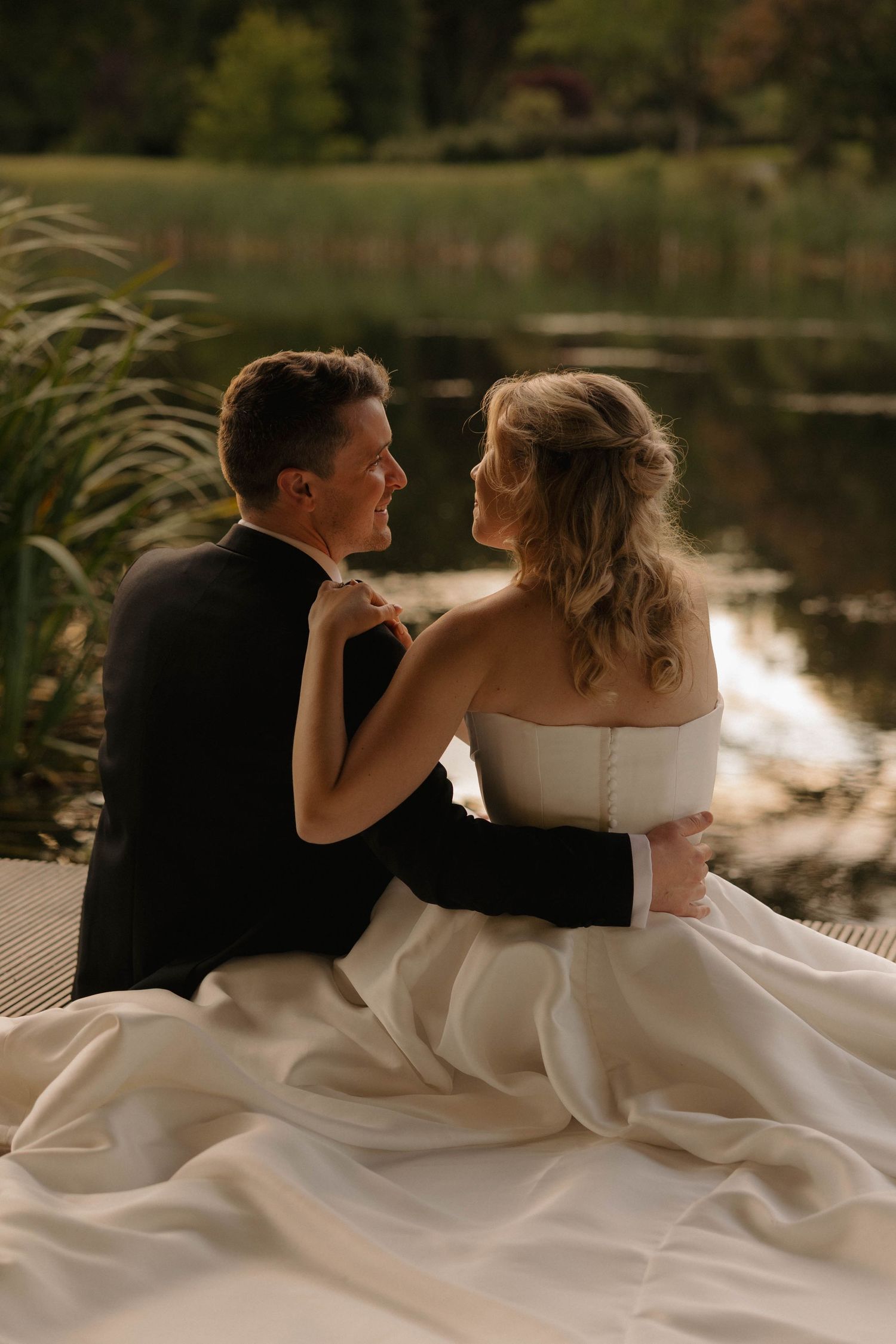 A romantic wedding couple sits by a serene lake at sunset, with the bride's flowing white dress cascading around them.
