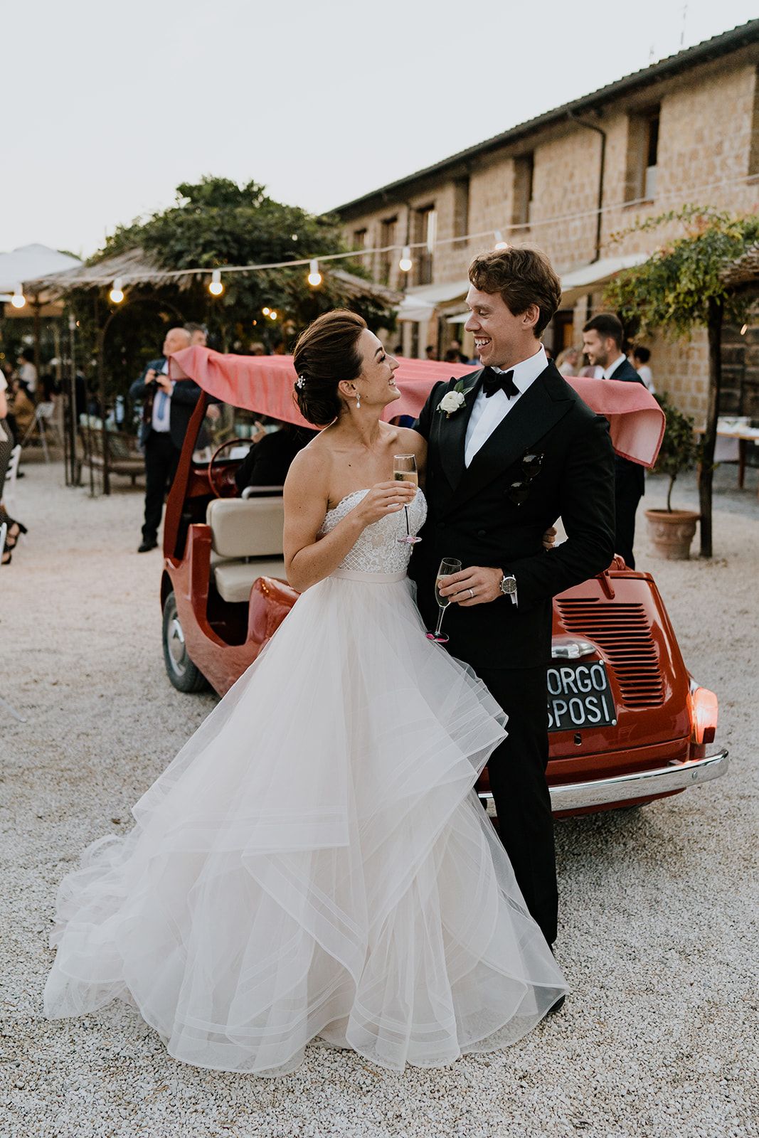 Wedding couple poses near a vintage red car in a rustic stone building courtyard.