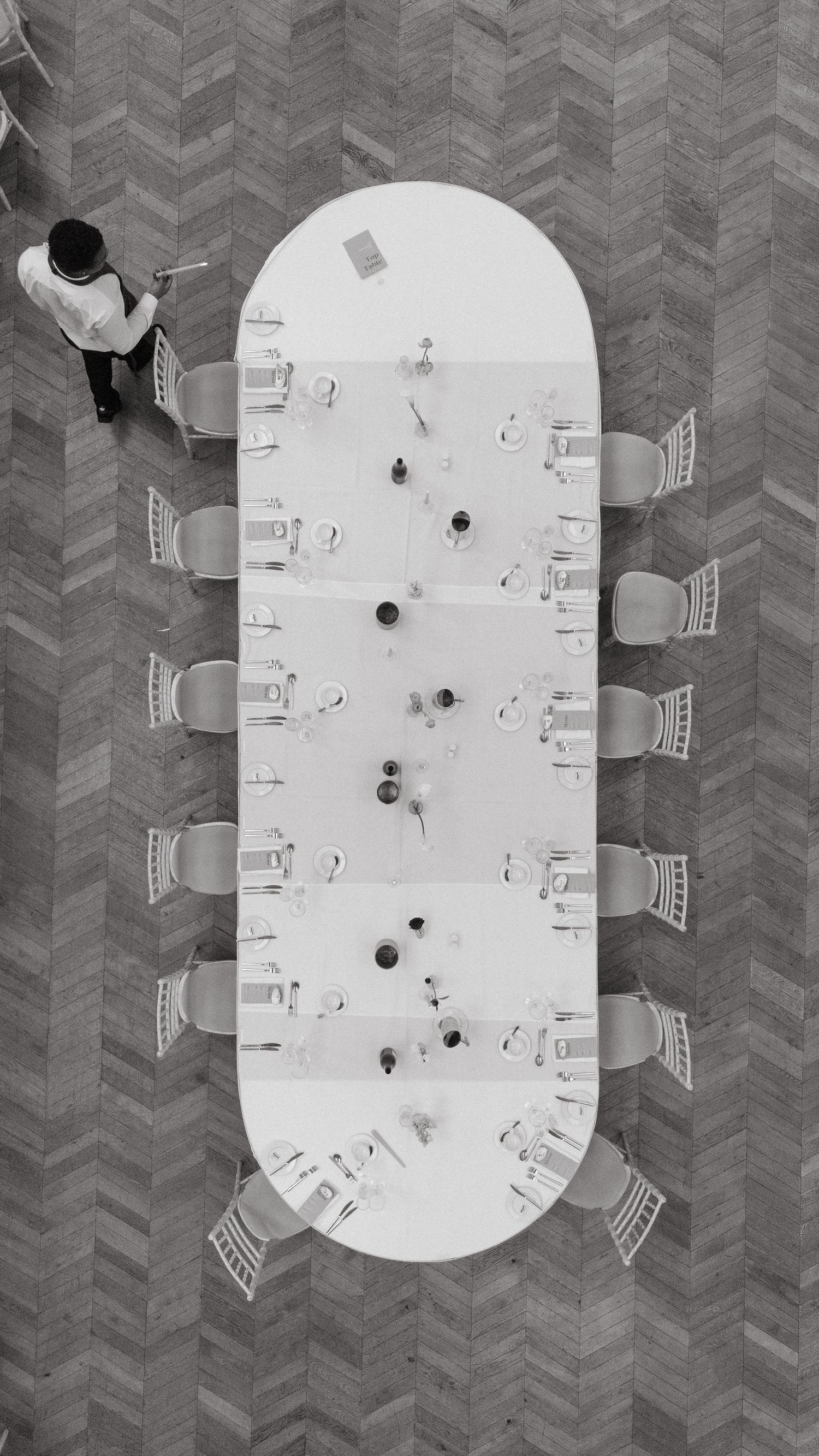 Aerial black and white view of a long oval conference table with chairs and place settings, with a person standing nearby.