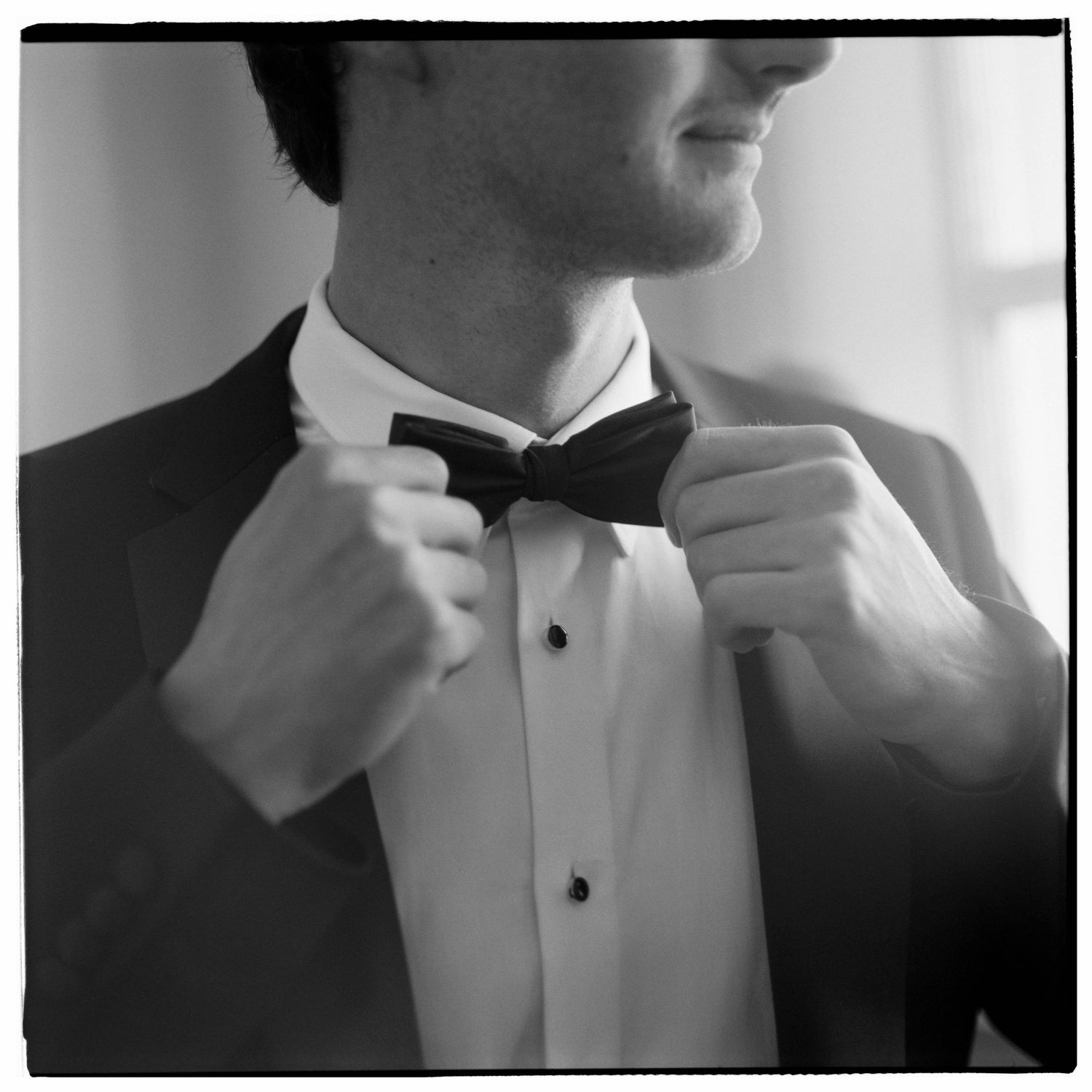 Groom in tuxedo adjusting black bow tie, black and white wedding photo.