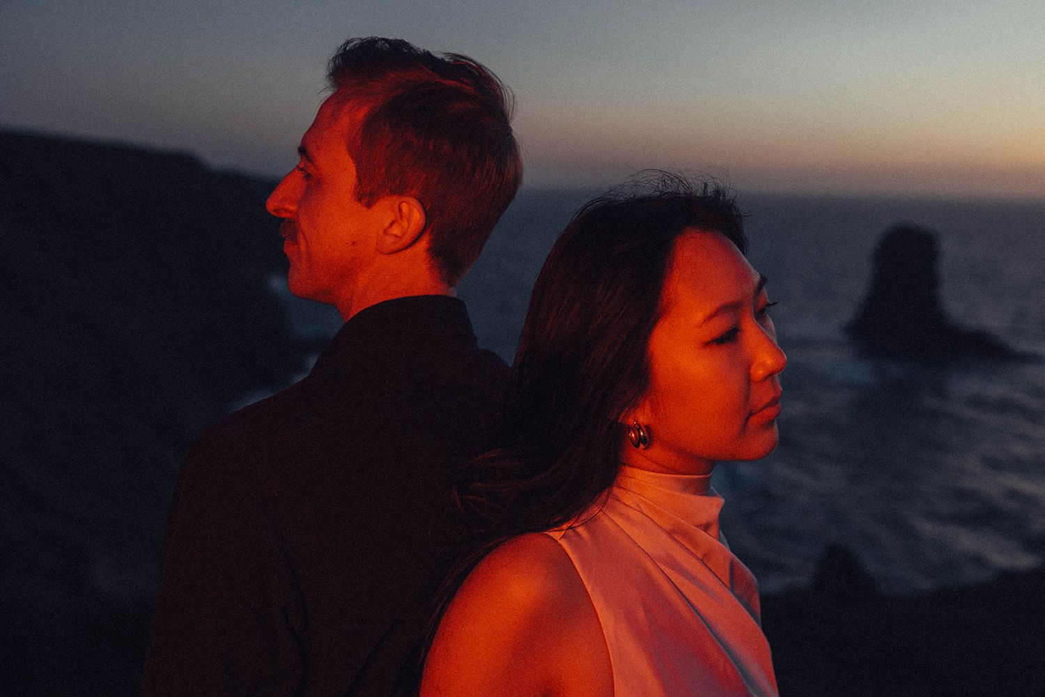 A couple stands back to back at sunset against a dramatic coastal backdrop with ocean stacks visible in the distance.
