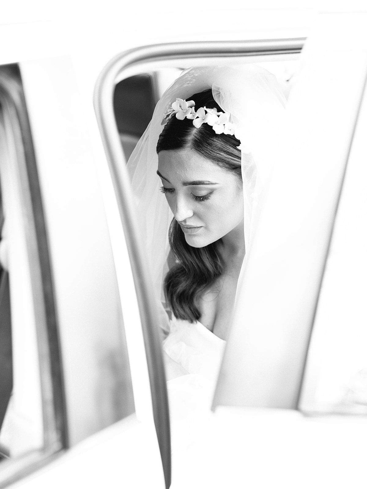 Black and white wedding photo showing a bride in white dress and delicate headpiece viewed through a car window frame.