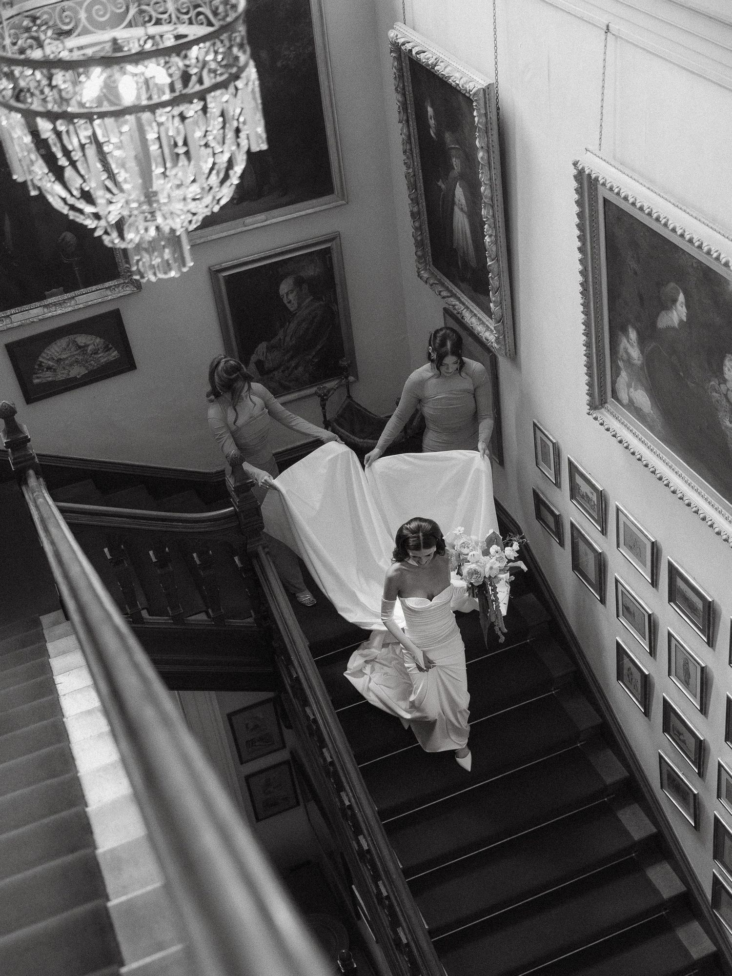 A black and white photo of an elegant staircase with ornate details and crystal chandelier in a historic interior setting.
