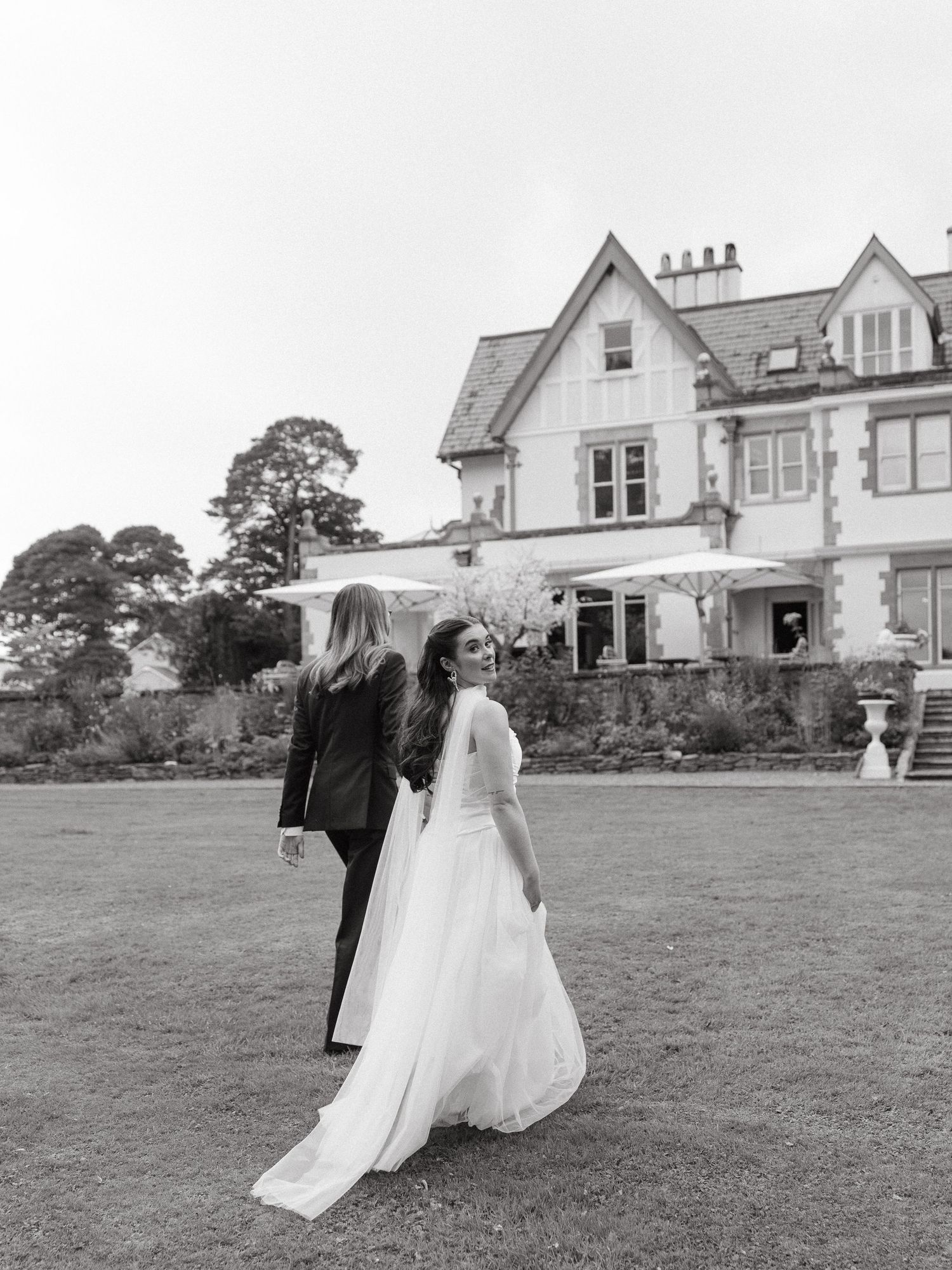 Black and white photo of a bride in a white gown and partner in a suit walking on a lawn before a grand manor house.