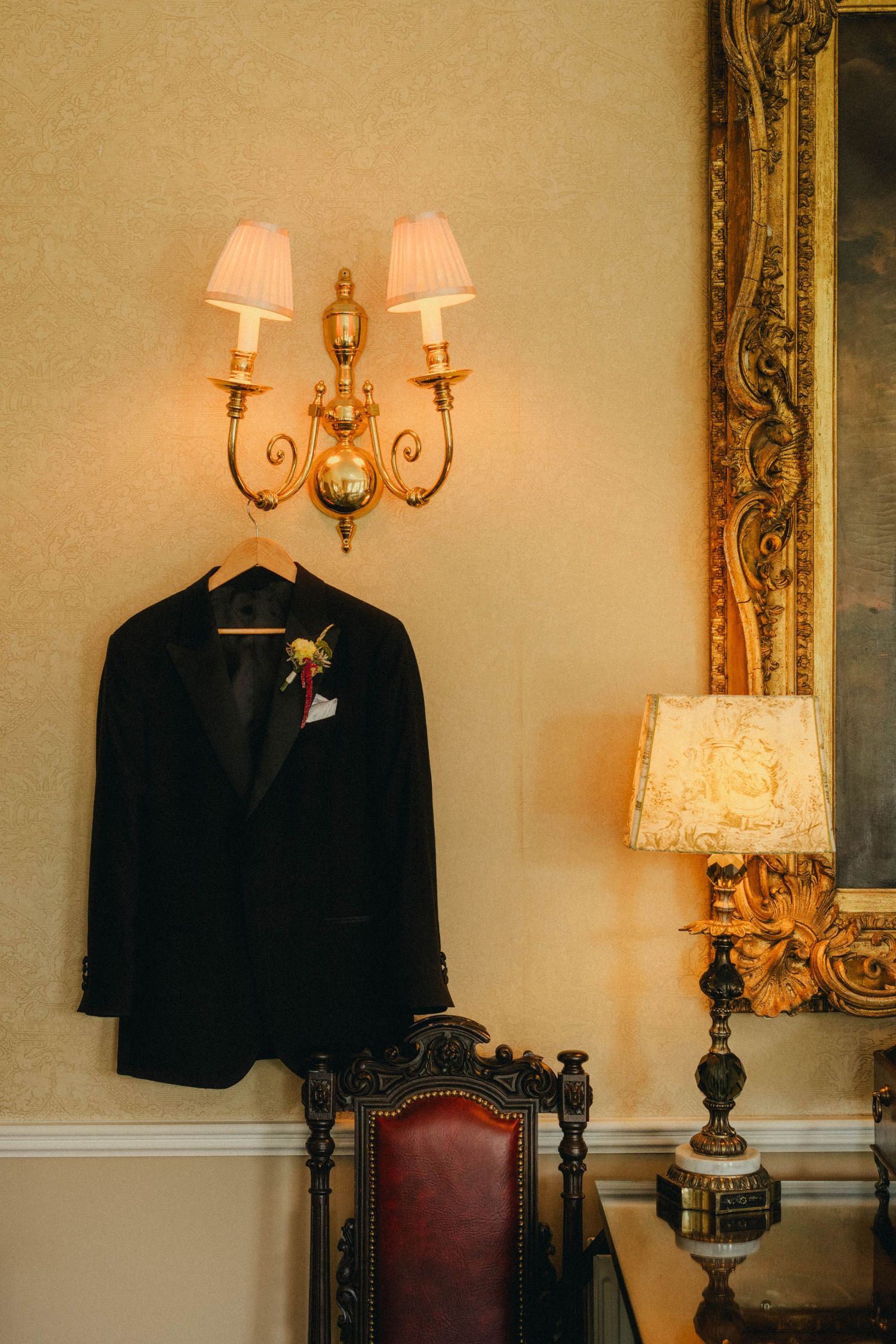 Black tuxedo jacket with boutonniere hanging on ornate gold wall sconce in elegant vintage room with gilded mirror.