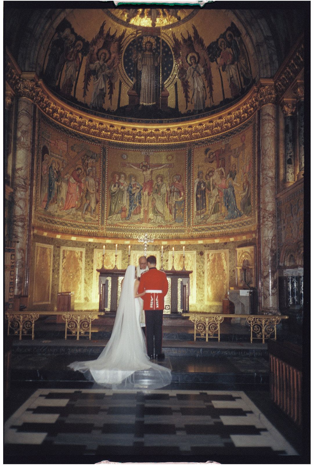 A bride and groom stand at the altar of an ornate church with golden mosaic artwork and religious murals in the apse.