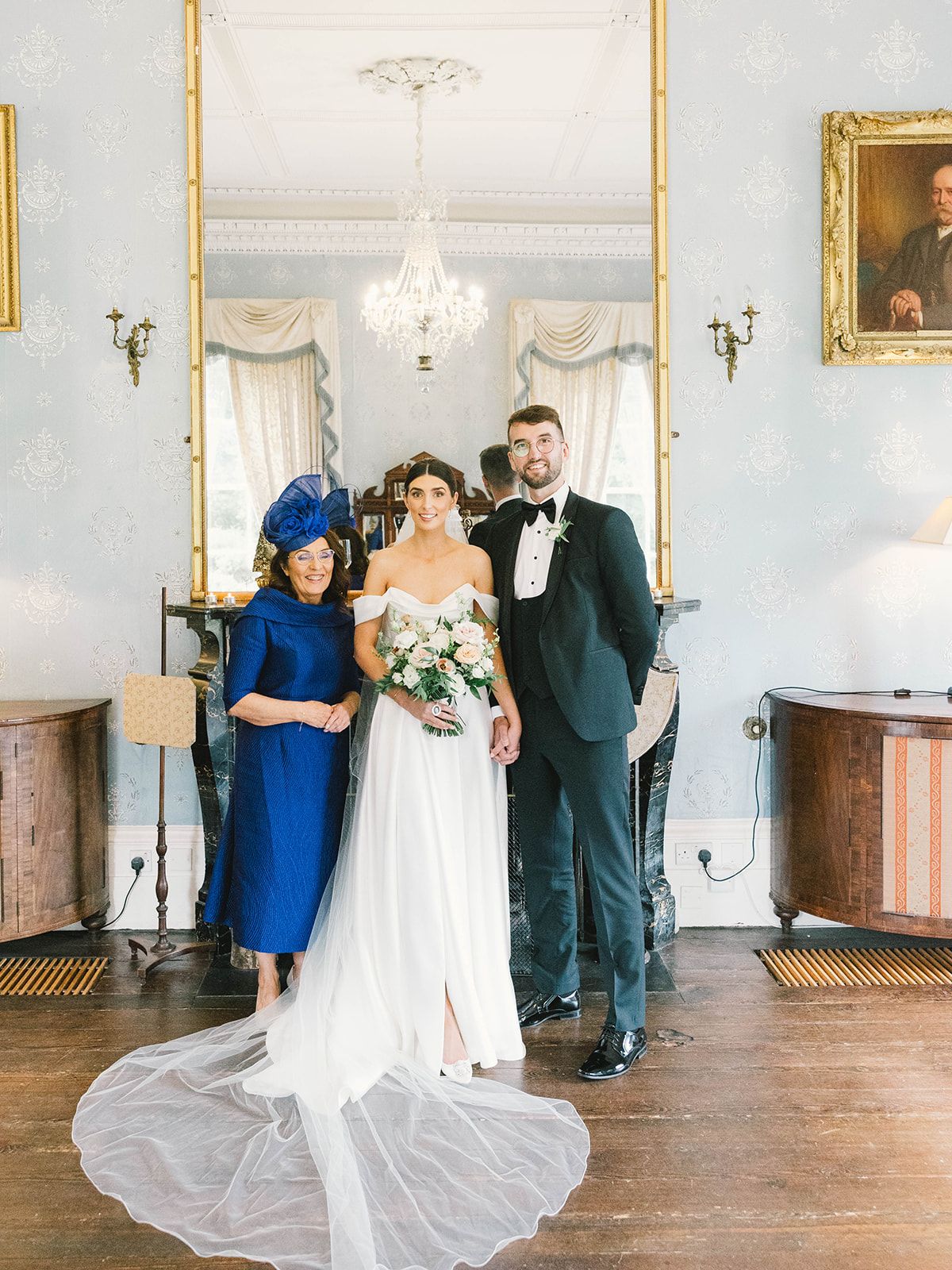 An elegant wedding photo in a stately room with chandelier, showing three people in formal attire posing together.