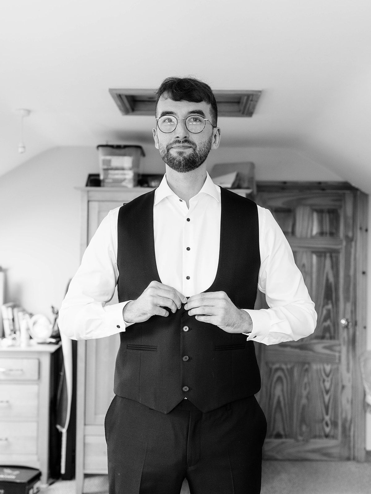 A person in formal attire with glasses and a beard buttons a black vest while wearing a white dress shirt.
