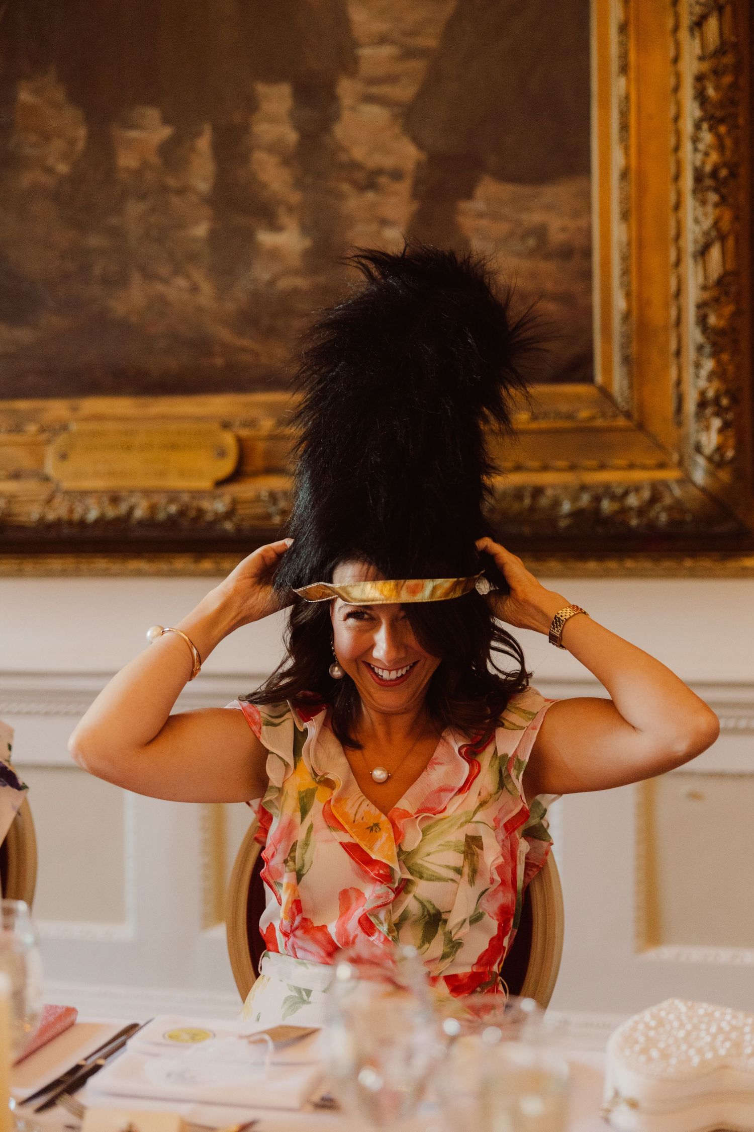 Wedding guest makes silly faces while joyfully adjusting a black feathered fascinator at an elegant reception table.