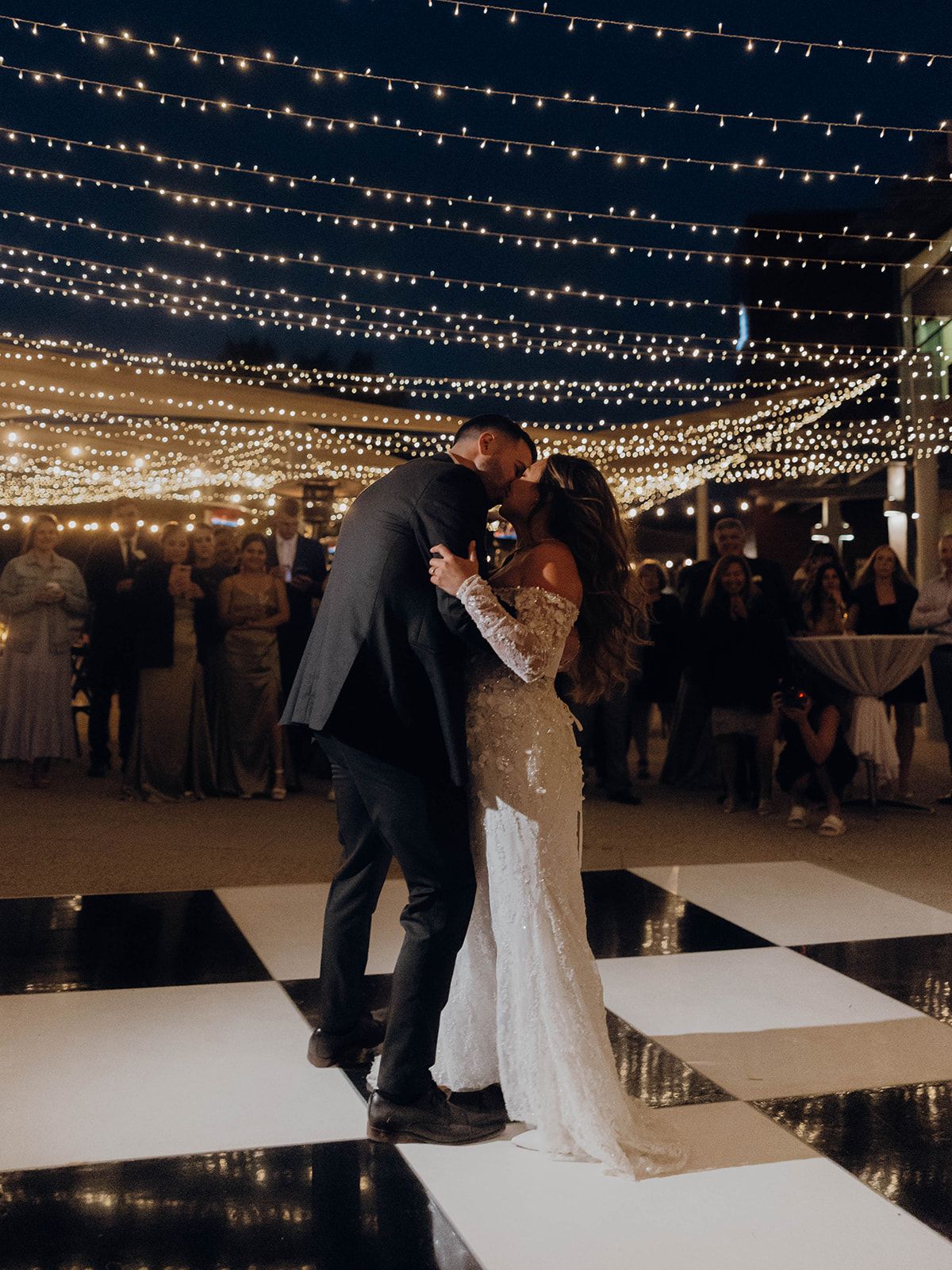 Romantic dance under twinkling string lights on a black and white checkered dance floor at an evening wedding reception.