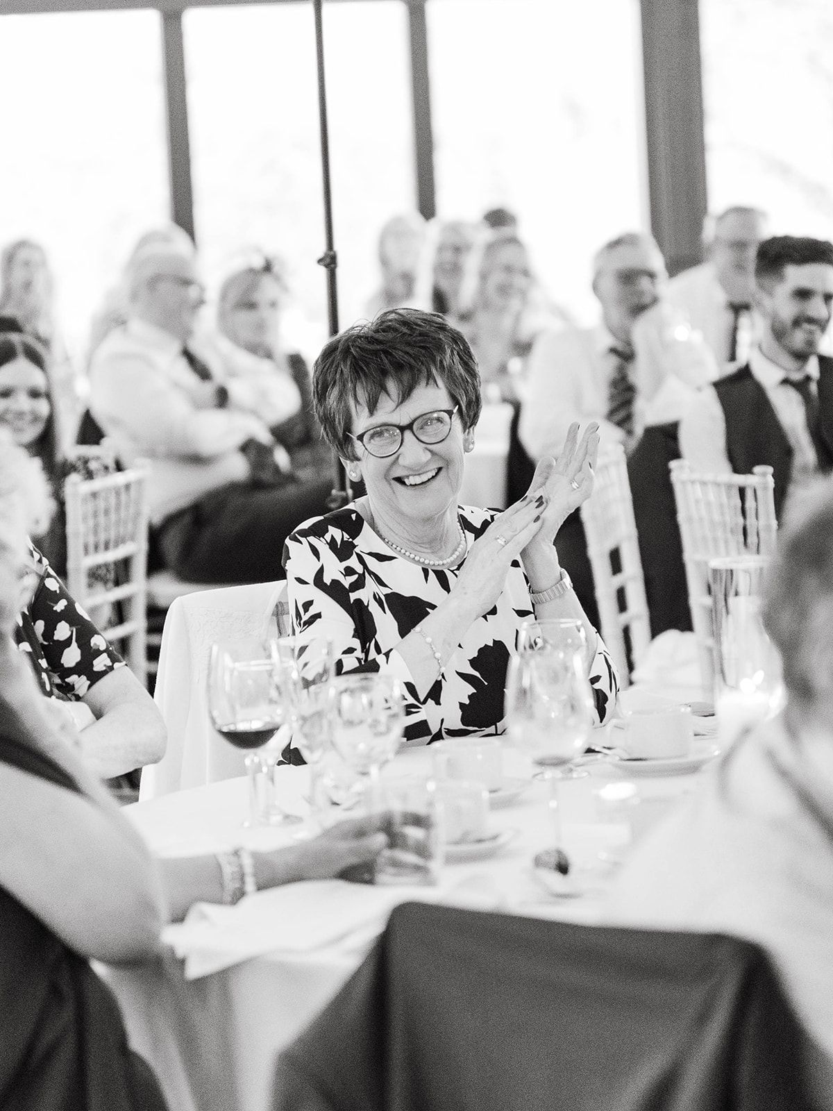 Black and white photograph of a wedding guest enjoying a reception dinner while seated at a table with wine glasses and decor.