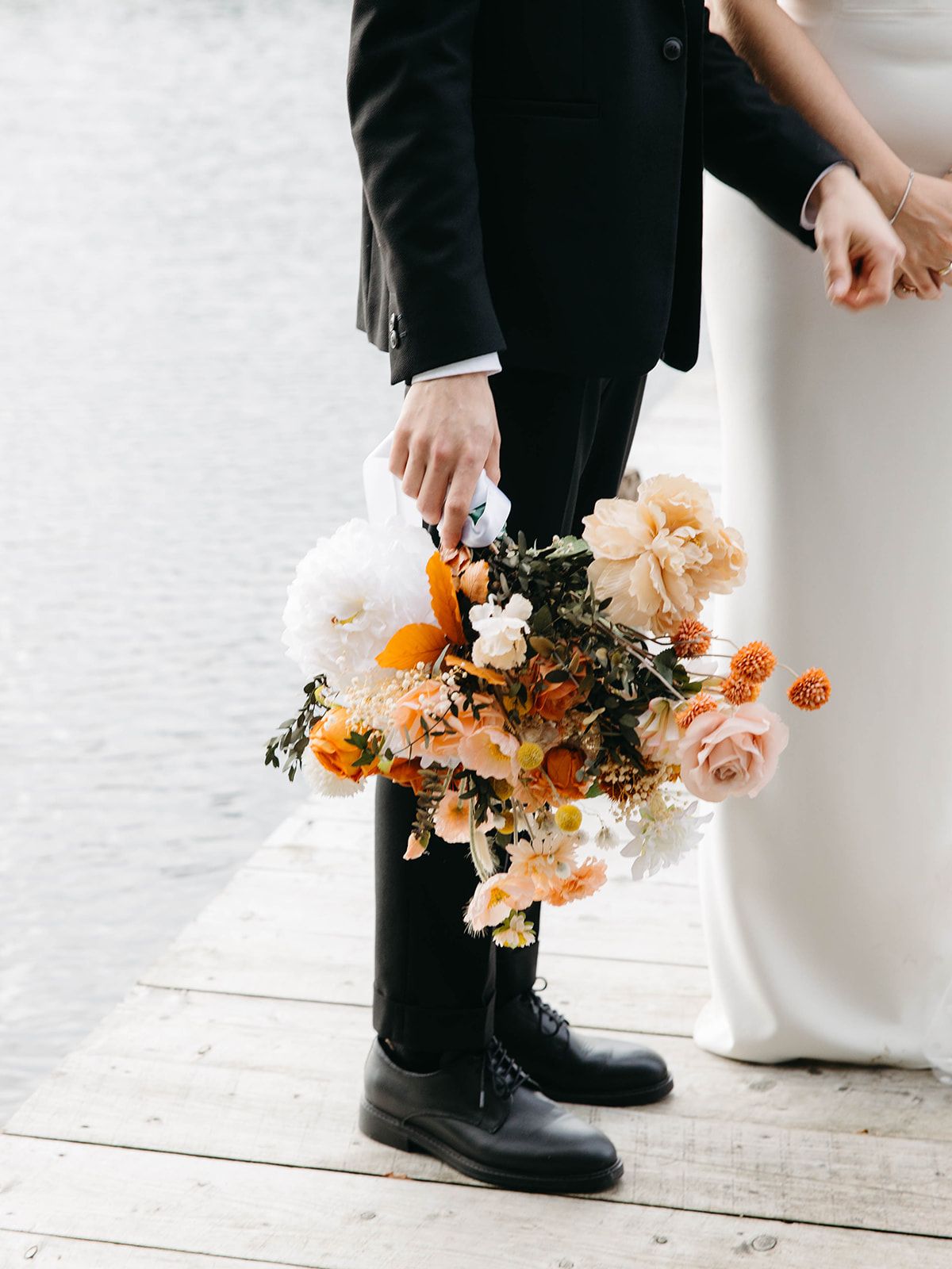 A wedding photo detail showing a person in black suit holding a peach and orange floral bouquet against a white background.