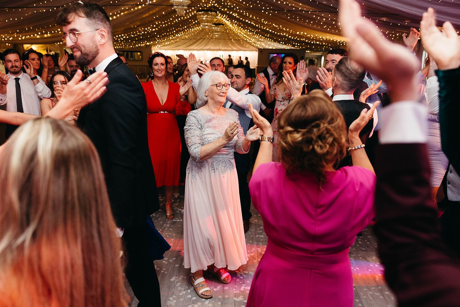 A lively wedding reception dance floor with guests in colorful dresses dancing under twinkling string lights.