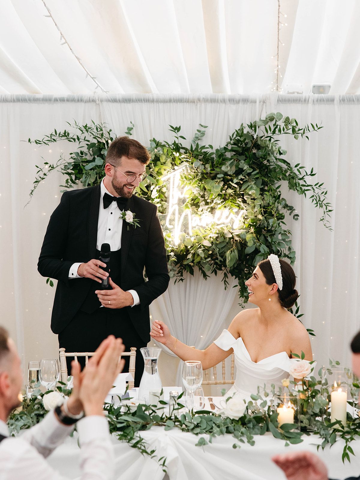 Elegant wedding reception scene with eucalyptus decor, white drapery, glowing candles and formal attire at head table.