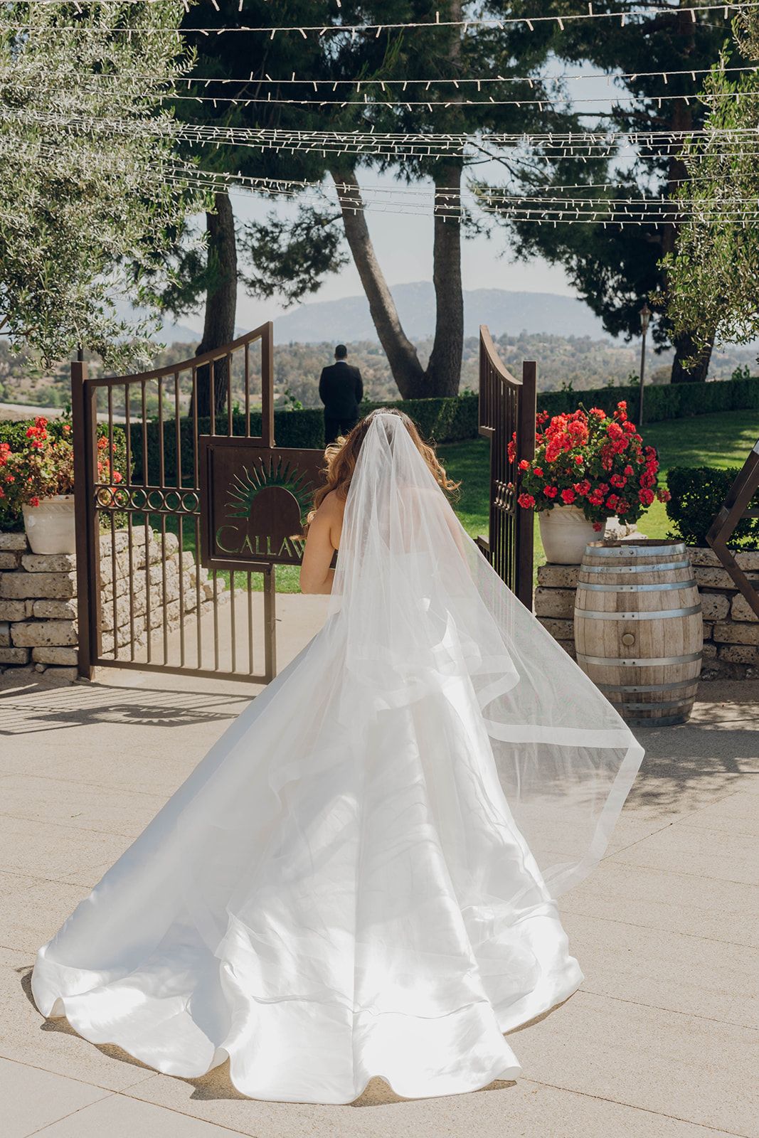 A flowing white wedding dress with a dramatic train photographed at an outdoor venue with string lights and stone walls.
