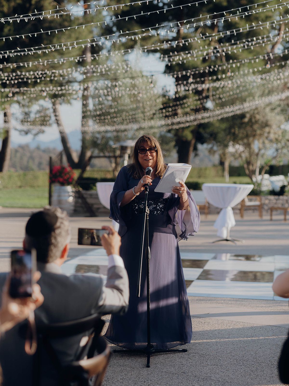 A person reads from papers at an outdoor event venue decorated with string lights overhead, while someone records on a phone.