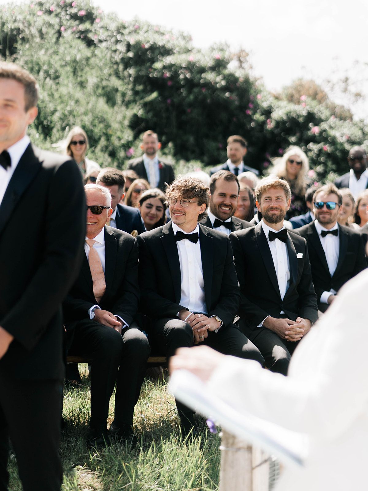 Groomsmen in black tuxedos seated during an outdoor wedding ceremony.