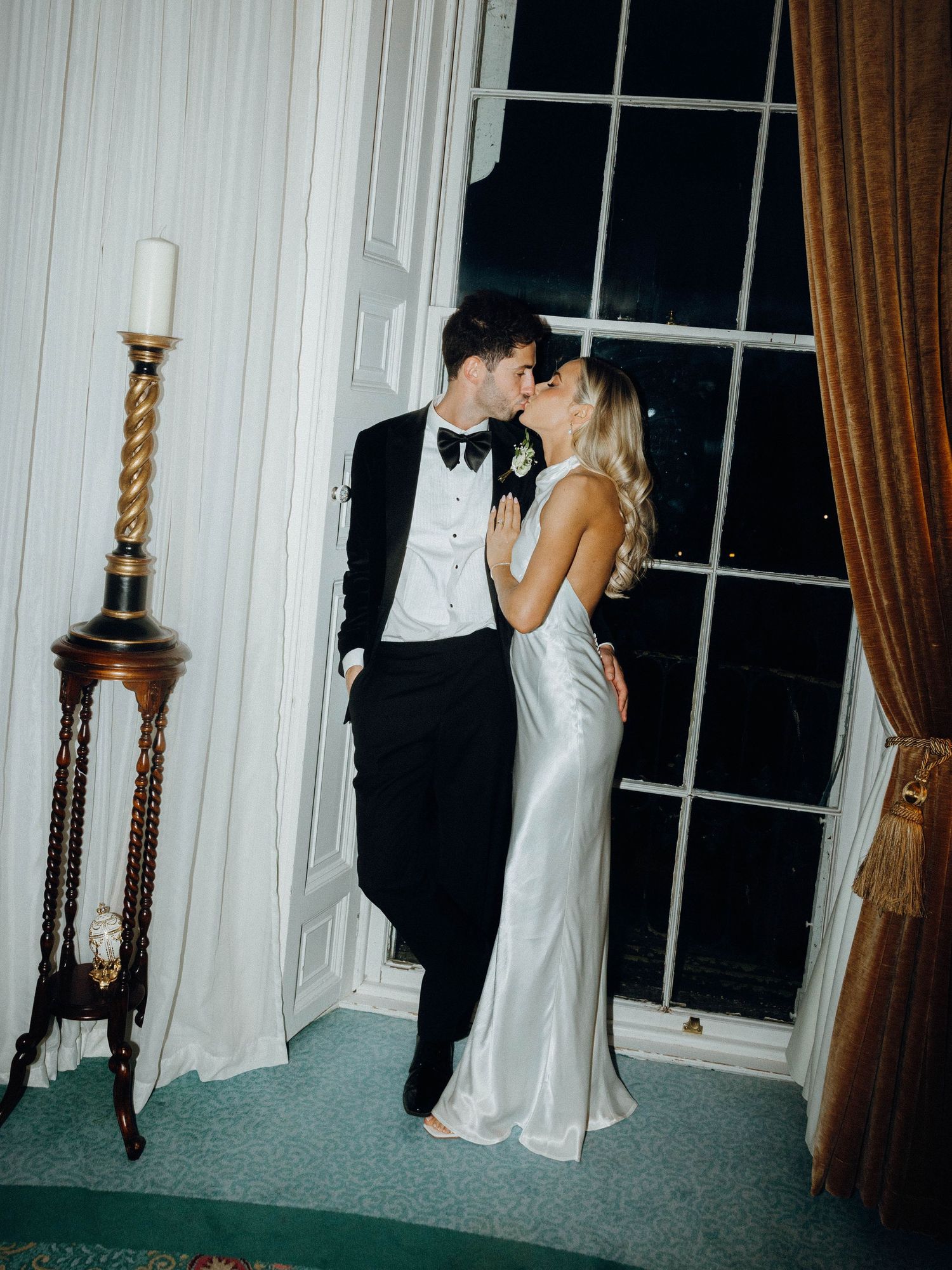 A couple in formal evening wear share a romantic moment by a tall window with an antique candlestick nearby.
