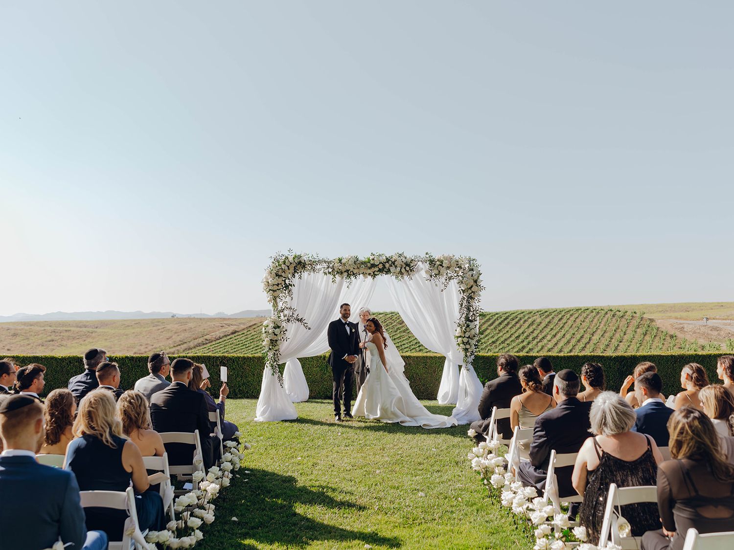 Outdoor wedding ceremony at a vineyard with white floral arch and guests seated on white chairs overlooking scenic hills.