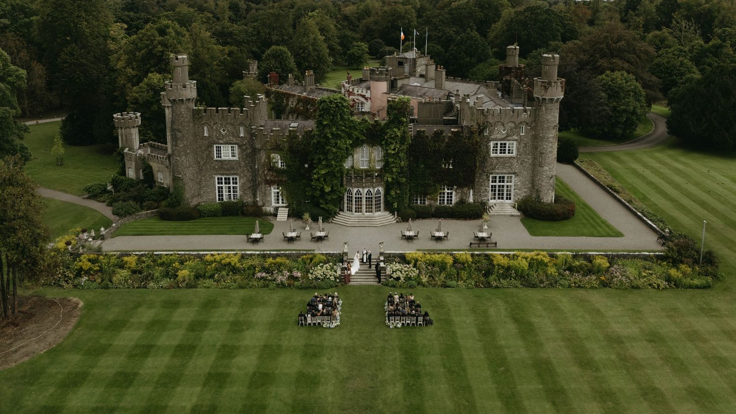 Aerial view of a stunning Irish castle estate with manicured lawns and wedding guests gathered outside.