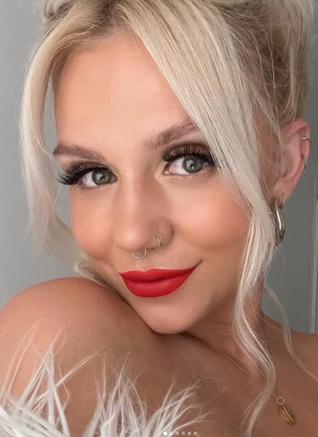 A close-up beauty portrait featuring platinum blonde hair, bold red lipstick, dramatic lashes and a nose piercing.