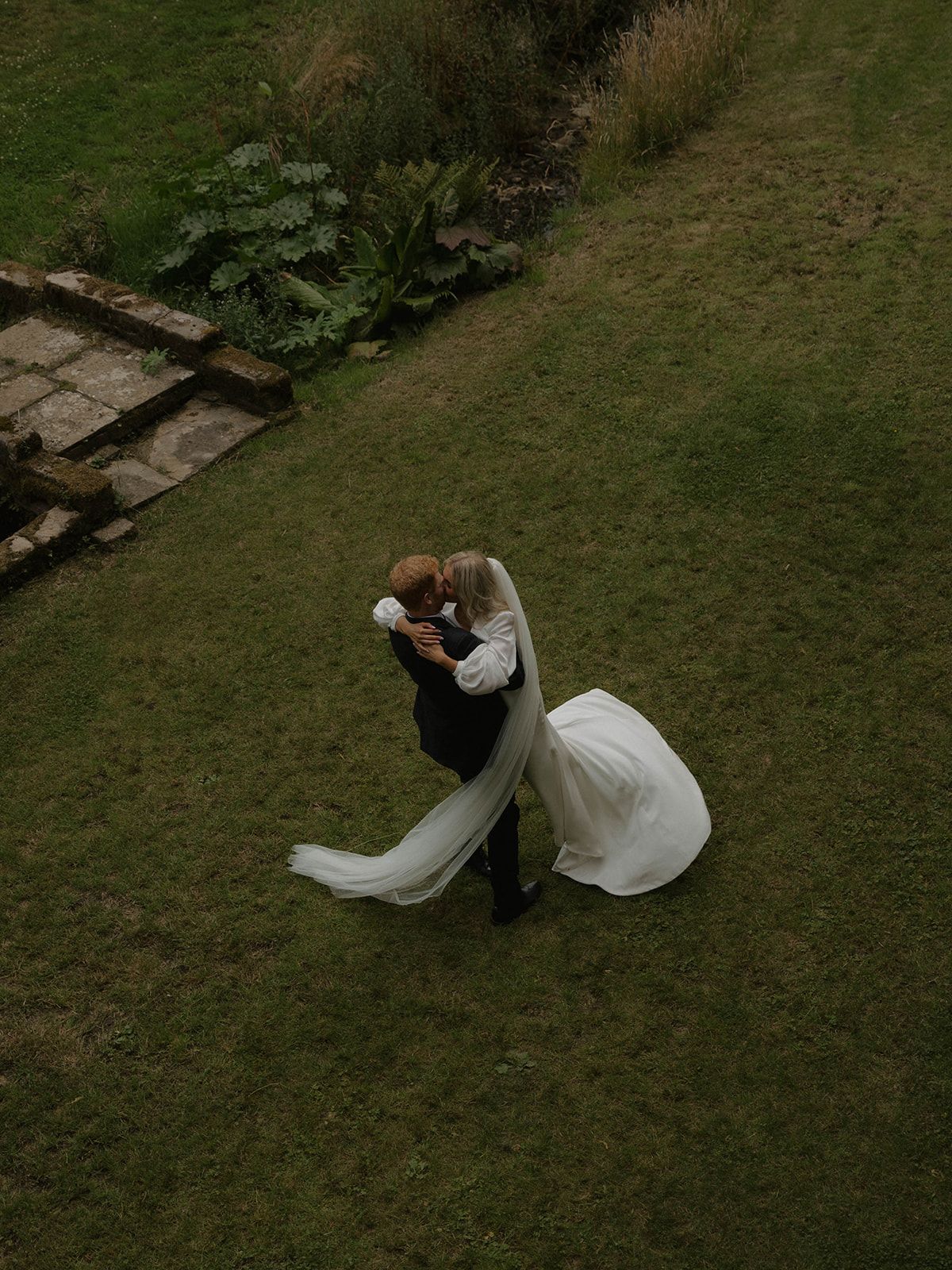 A couple in formal attire performs an elegant dance dip on a grassy lawn during what appears to be a wedding photoshoot.