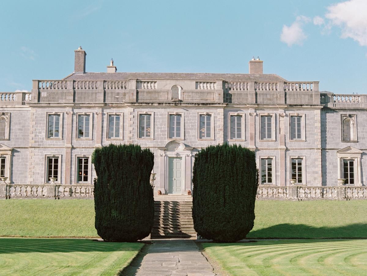 Historic stone castle with manicured lawns and tall hedges stands against a bright blue sky.