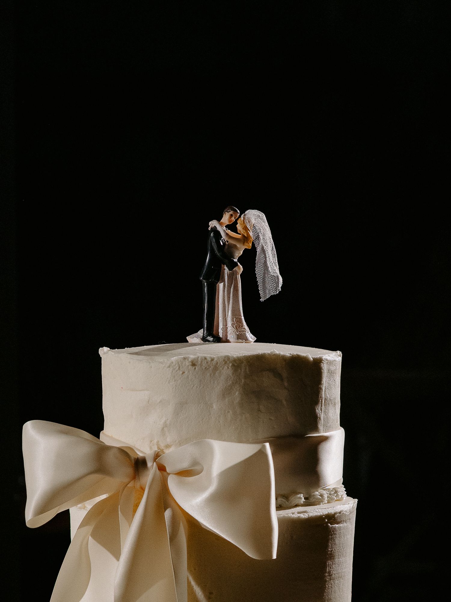 Elegant white wedding cake with satin bow and classic bride and groom figurine topper on dark background.