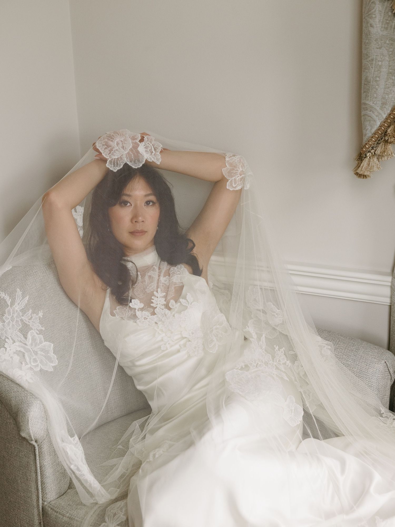 A bride in a white lace wedding dress and veil sits gracefully on a chair with arms raised.