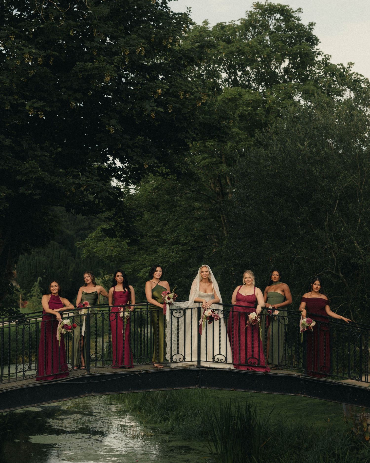 Bride in white gown with bridesmaids in red and green dresses posing on a bridge surrounded by lush greenery.